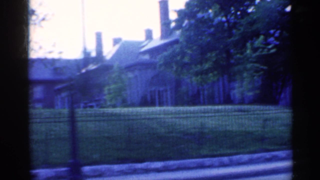 1959 Indiana home-movie view of a stately residence with chimneys, shaded trees, lawn, and roadside iron fencing.