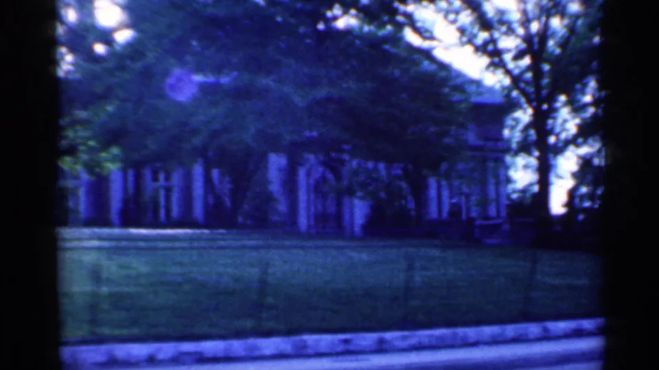 Blue-tinted 1959 Indiana 8mm frame of a shaded residential facade, broad lawn, trees, and low iron fence.