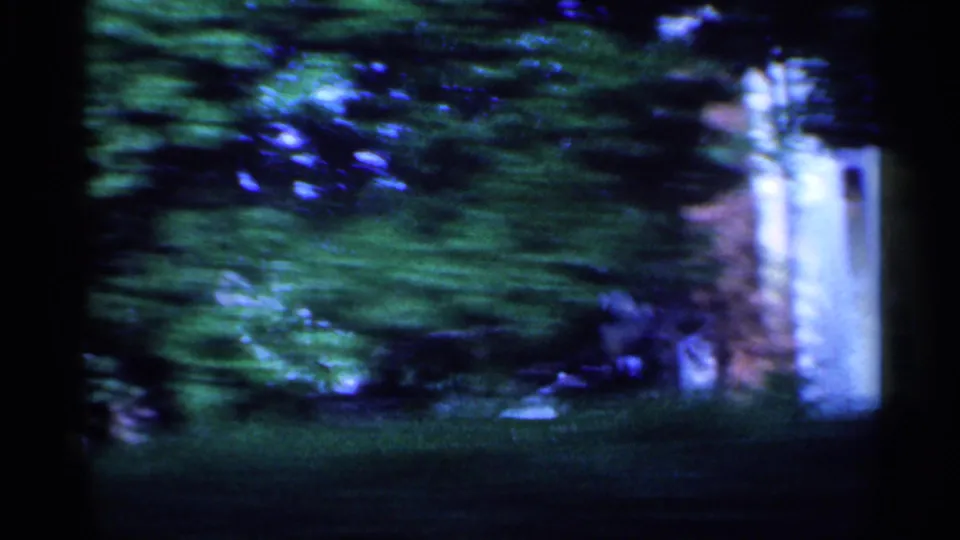 Motion-blurred 1959 Indiana home-movie frame showing greenery, a pale wall, and shaded residential grounds from the road.
