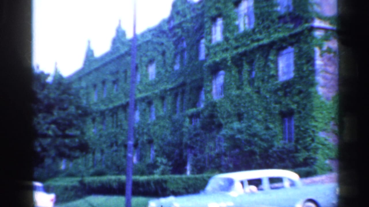 1959 Indiana 8mm frame showing an ivy-covered building facade, clipped hedges, trees, and a pale mid-century car near the curb.