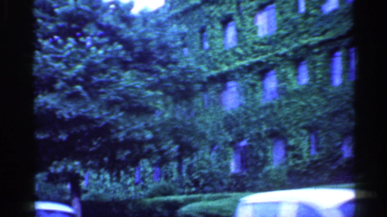 Blue-tinted 1959 Indiana home-movie frame with trees, an ivy-covered building, hedges, and part of a pale classic car.