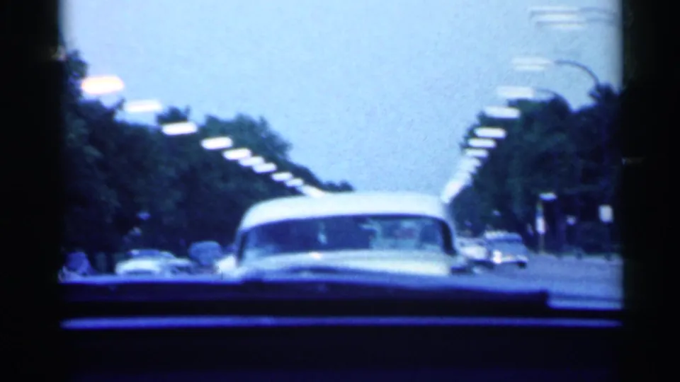 Passenger-view 1959 Indiana 8mm frame following a pale classic car along a tree-lined road with street lamps and parked vehicles.