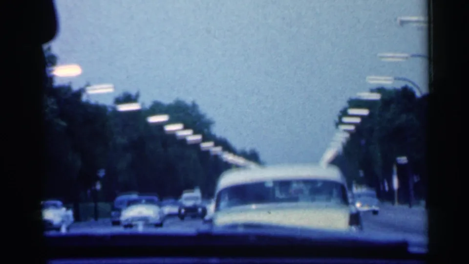 1959 Indiana driving view from inside a car, with a pale mid-century sedan ahead, rows of trees, street lamps, and distant traffic.
