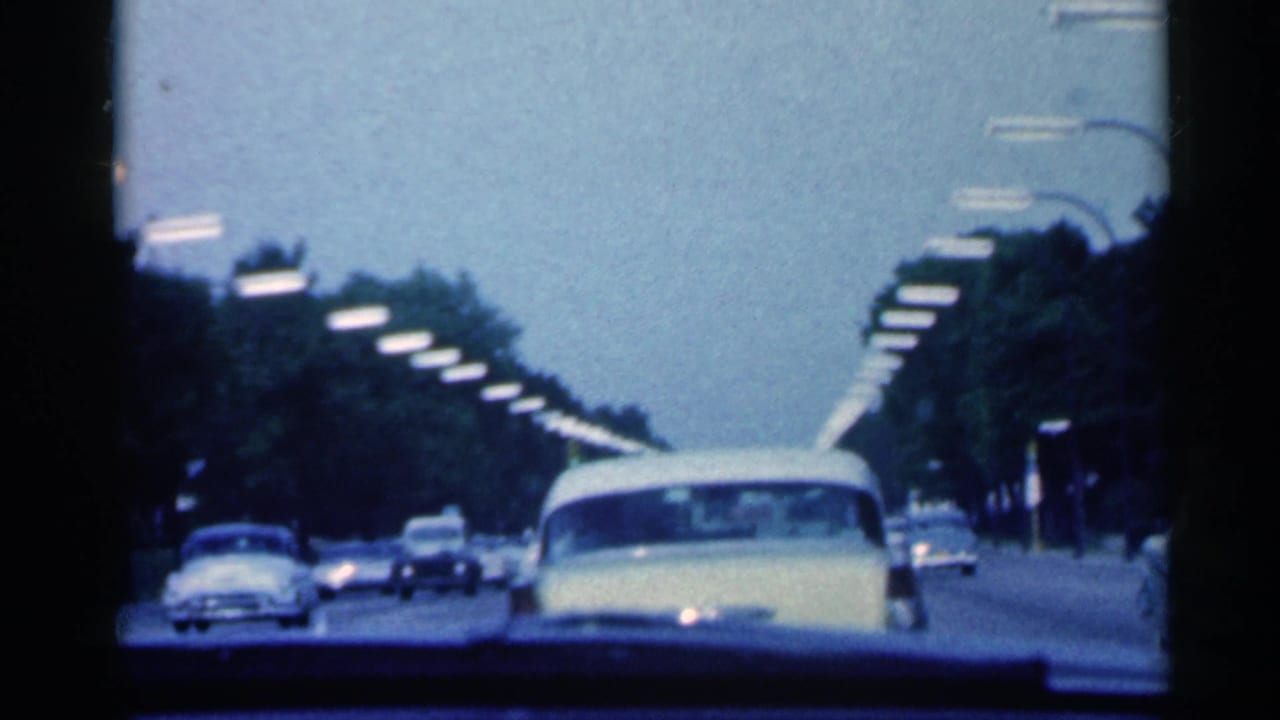 1959 Indiana 8mm road view from inside a moving car, with a pale yellow sedan ahead and traffic under rows of streetlights.