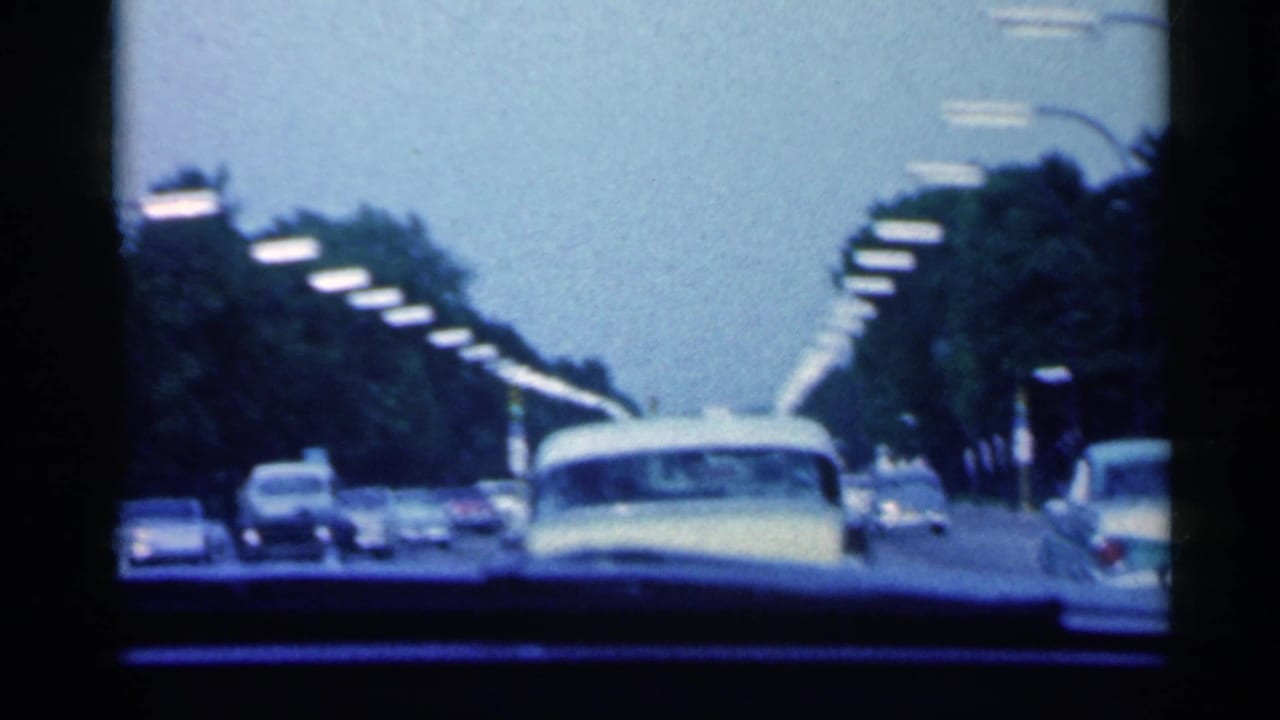 A 1959 Indiana home-movie frame shows several cars ahead on a tree-lined roadway, with dark dashboard and windshield edges visible.