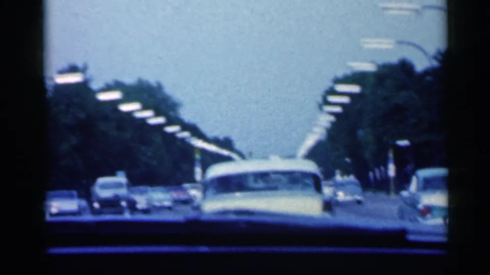 A 1959 Indiana home-movie frame shows several cars ahead on a tree-lined roadway, with dark dashboard and windshield edges visible.