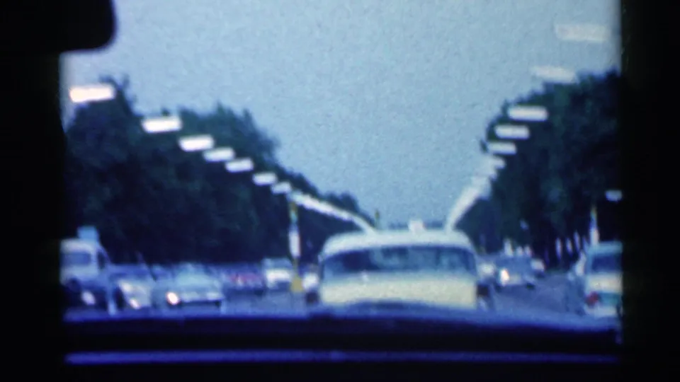 From inside a moving car, the 8mm frame follows a pale yellow sedan among late-1950s traffic on a tree-lined Indiana road.