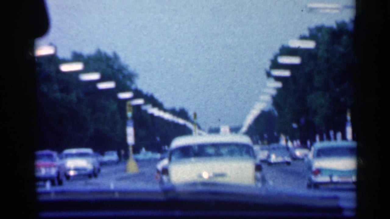 1959 Indiana home-movie view through a windshield as cars drive along a broad tree-lined road with streetlights.