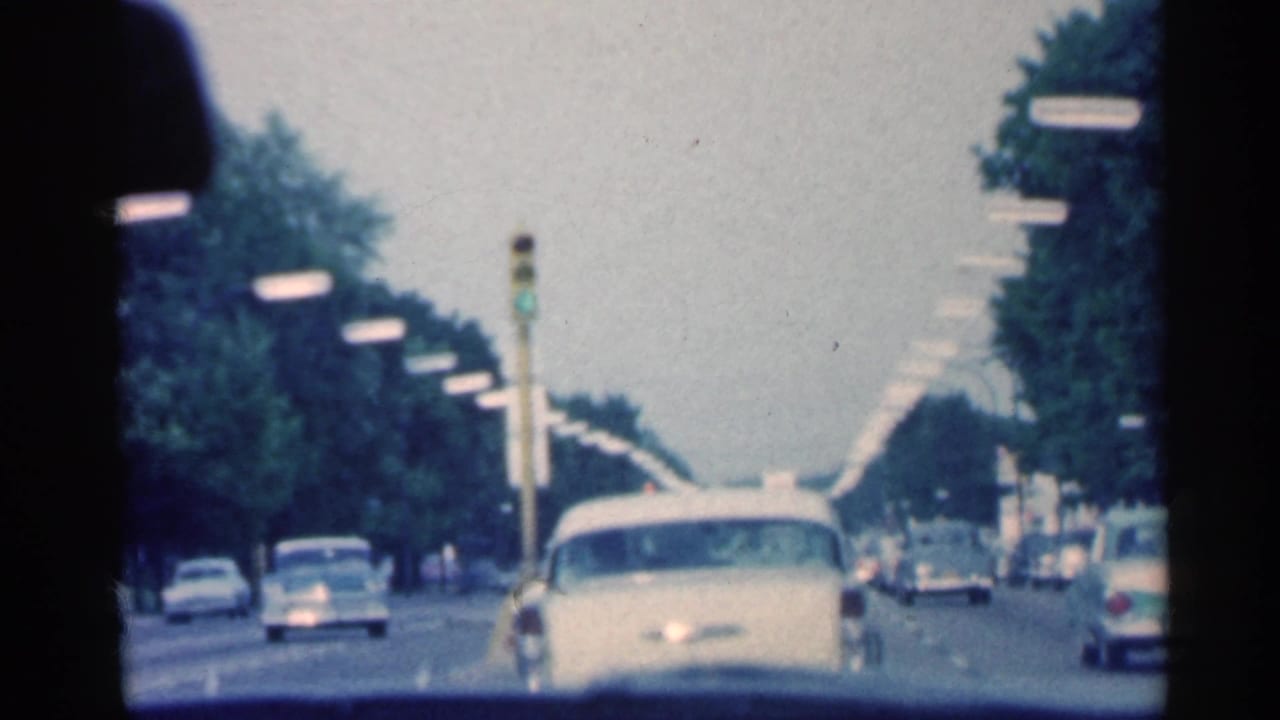 A white late-1950s sedan is seen ahead on an Indiana roadway, with traffic signals and repeated streetlights in view.