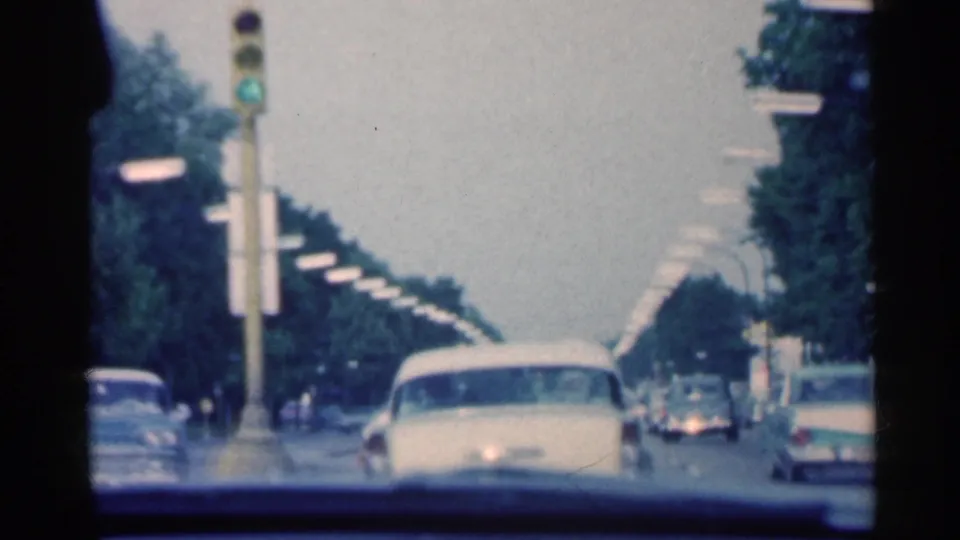 Windshield view of an Indiana street in 1959, with cars moving beneath a green traffic signal and rows of roadside lights.