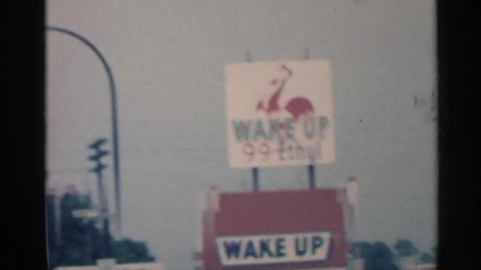 1959 Indiana roadside view with a red and white Wake Up 99 Ethyl sign and rooster logo seen from a moving 8mm camera.