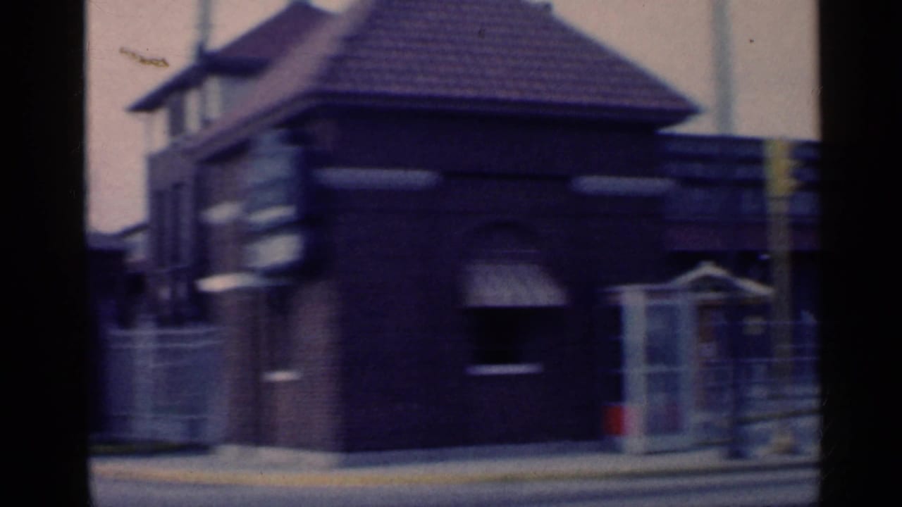 A brown brick building with a dark tiled roof passes the camera during the 1959 Indiana roadside home-movie shot.
