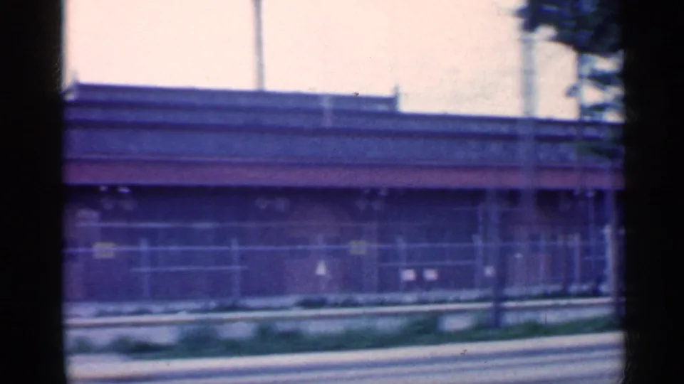 A long low commercial frontage and fence slide past the 8mm camera, with poles and roofline visible in Indiana roadside footage.