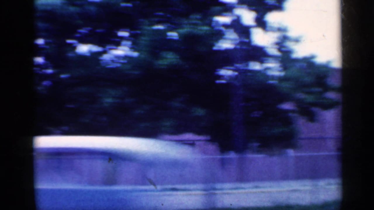 A moving 8mm view in Indiana, 1959, with trees and a low roadside wall partially obscuring buildings beyond.