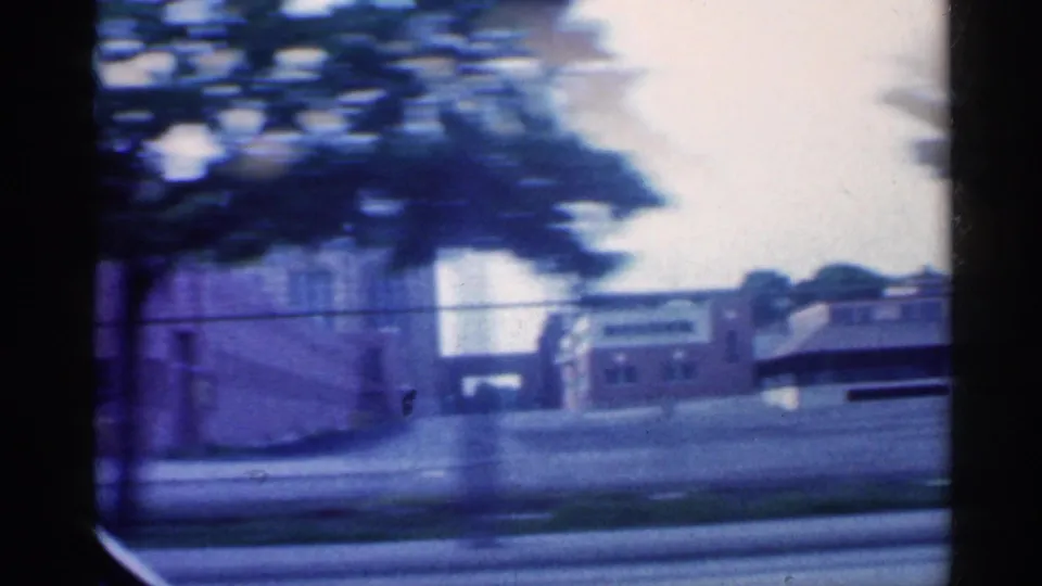 1959 Indiana street view from a moving vehicle, with brick buildings, trees, and a broad roadway softened by motion blur.