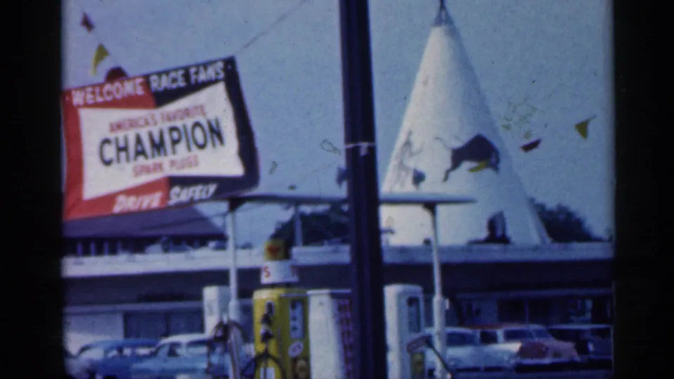 1959 Indiana roadside frame showing a Champion Spark Plugs sign, parked cars, pennants, and a cone-shaped structure behind it.