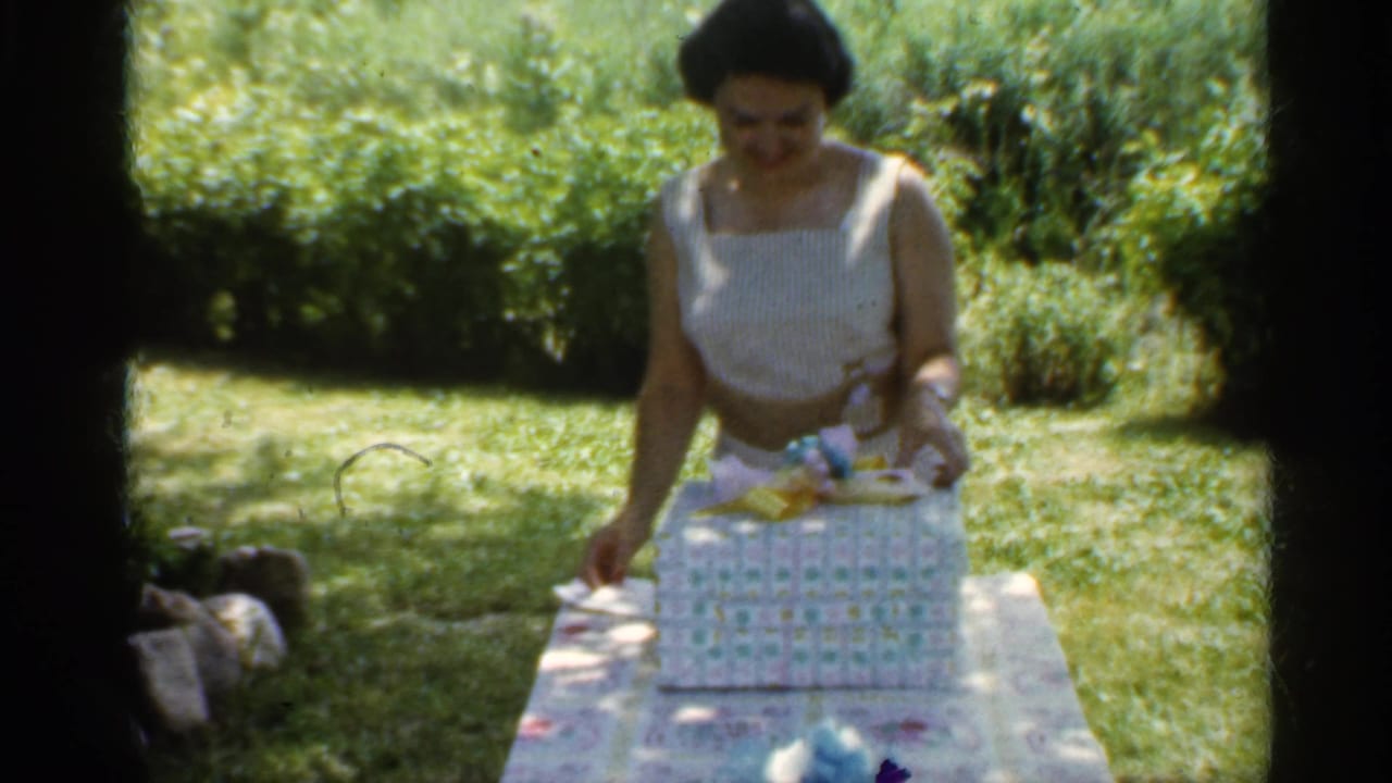 Woman in a white sleeveless dress stands at a garden table in Indiana, opening a wrapped present in 1959 footage.