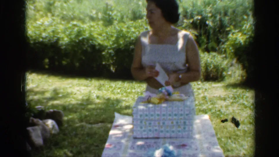 Woman in a white sleeveless dress holds wrapping or an envelope beside the present on a garden table in Indiana, 1959.