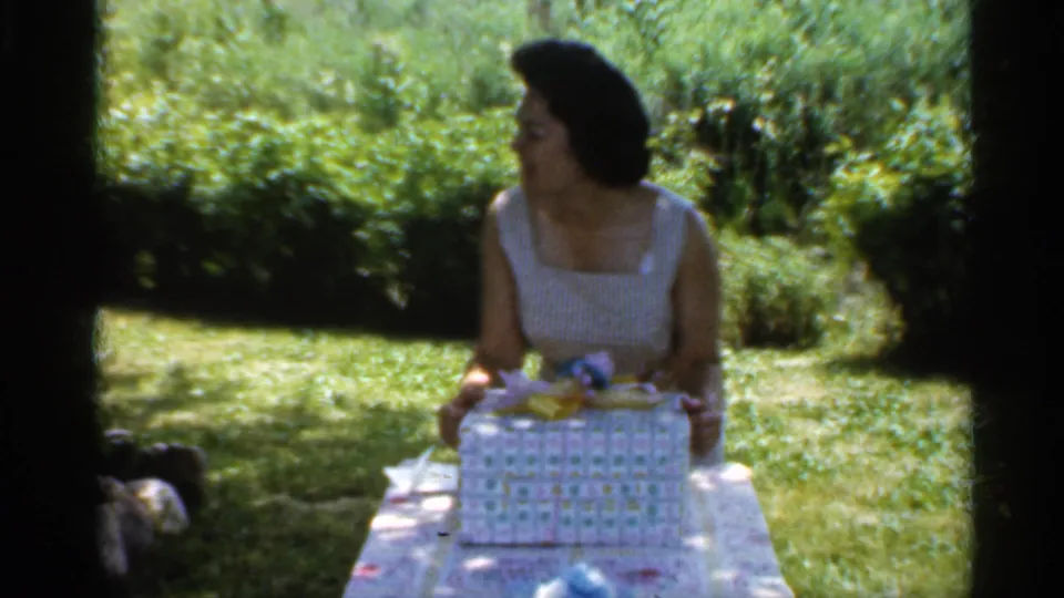 Woman at a sunlit garden table looks aside with the opened gift box in front of her, from Indiana 1959 home-movie footage.