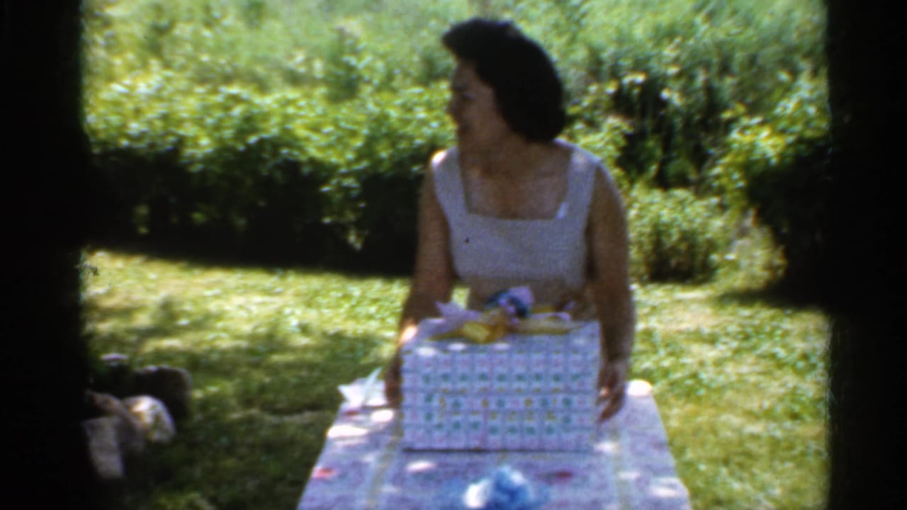 In 1959 Indiana home-movie footage, a woman in a white sleeveless dress sits at a backyard table with a wrapped birthday gift.
