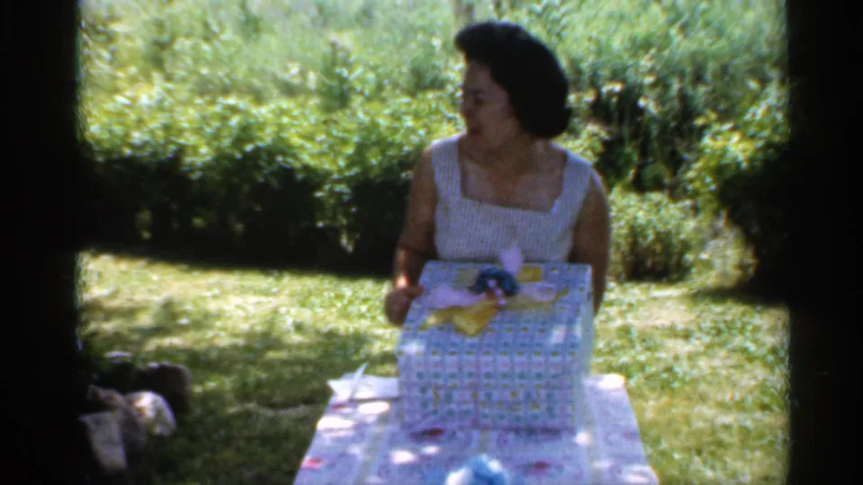 A woman seated at a table in 1959 Indiana home-movie footage holds a patterned gift box in a bright backyard setting.