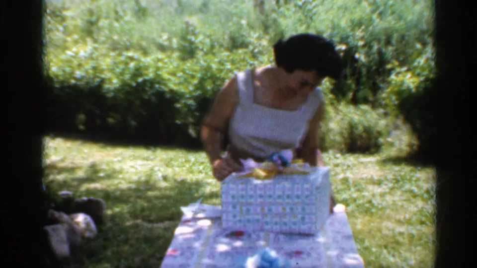 The woman leans toward the wrapped birthday gift on a backyard table, with lawn and shrubs visible behind her.