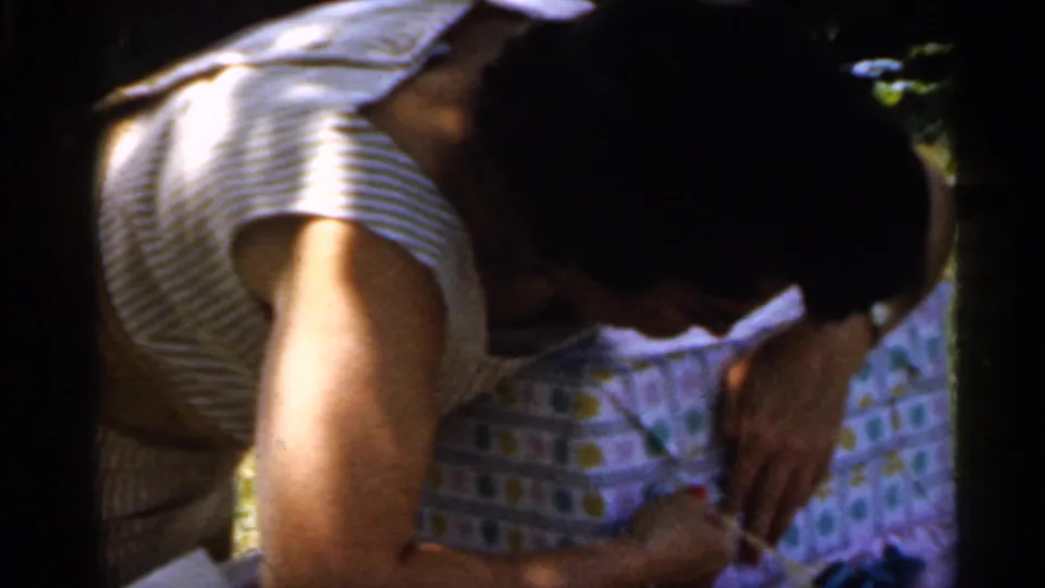 In 1959 Indiana home-movie footage, a woman in a striped sleeveless top leans over a wrapped birthday present outdoors.