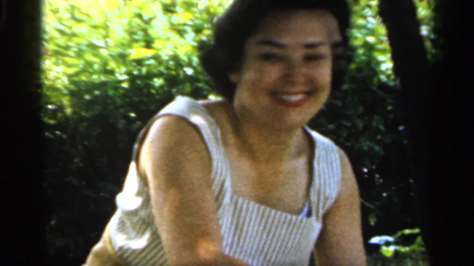 A smiling woman faces the home-movie camera on a leafy Indiana lawn, with soft 8mm grain and bright outdoor light.