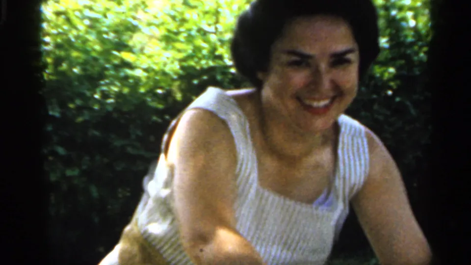 The woman leans forward and smiles on the grassy lawn, framed closely in late-1950s Indiana 8mm home footage.
