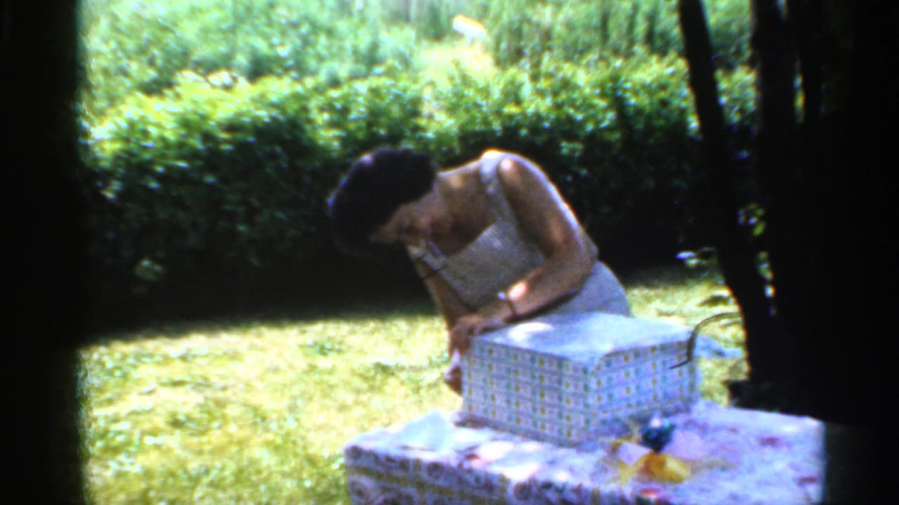 Adult woman in a white sleeveless dress leans over a gift box on a garden table.