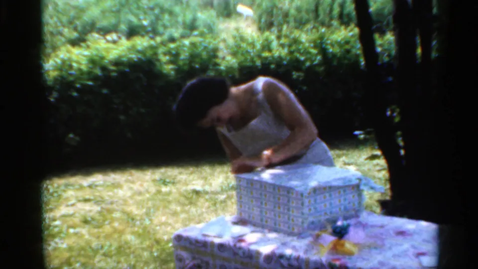 In a sunlit Indiana garden, the woman continues opening the gift box on a patterned tablecloth.