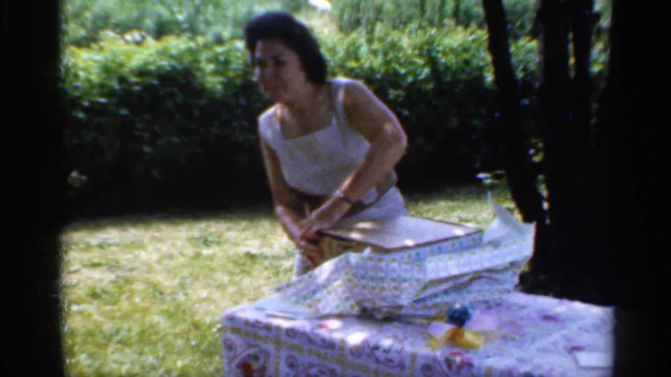 The woman lifts tissue or wrapping from the gift box during an outdoor 1959 Indiana home-movie celebration.