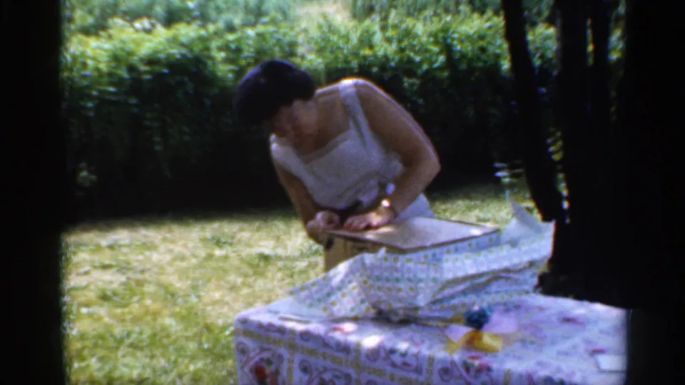 The woman bends over the opened present in the garden, framed by lawn, hedges, and tree shade in 1959 Indiana footage.