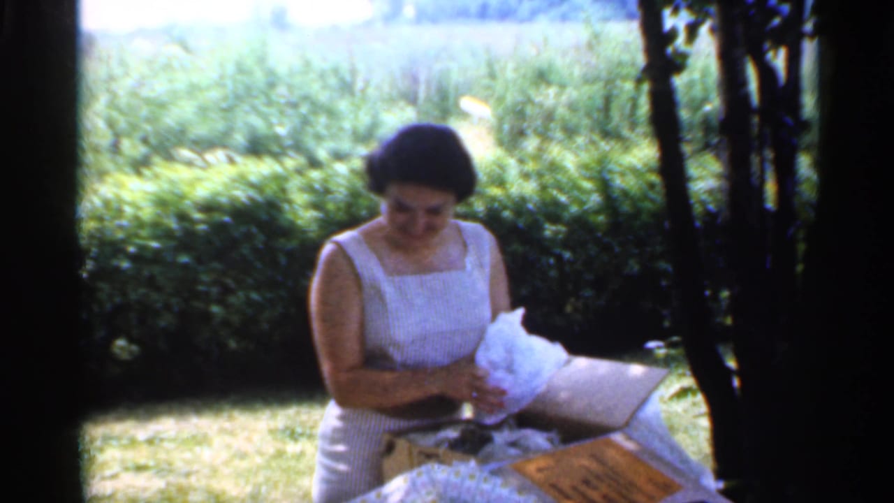 The woman in a light dress looks down at tissue wrapping beside an open box, with grass, hedge, and trees behind her in Indiana footage from 1959.
