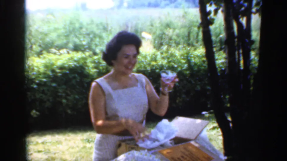 In the garden scene, the woman smiles toward the camera while raising a small glass object from tissue paper on the table.