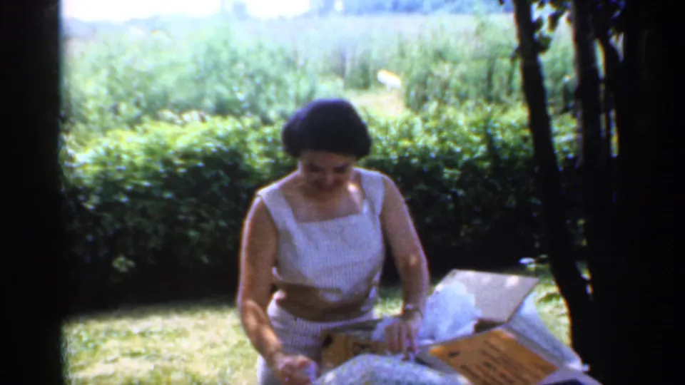 The woman leans over the outdoor table after unwrapping the item, with the open cardboard box, tissue, lawn, hedge, and tree shade visible.