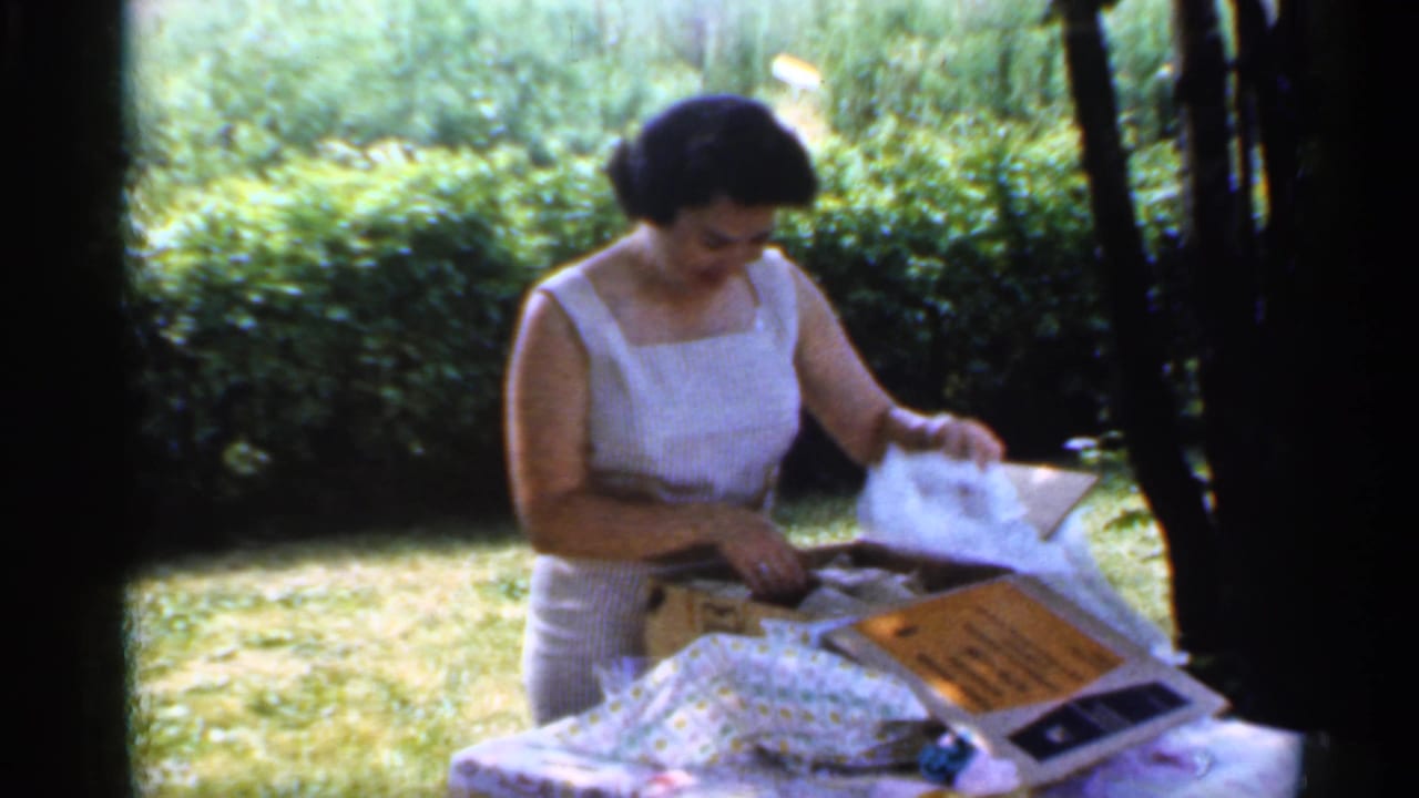 Woman in a white sleeveless dress opening gifts on a backyard table.