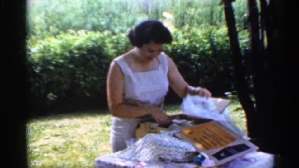 1959 Indiana home-movie frame of a woman in a white sleeveless dress opening wrapped gifts outdoors beside hedges.