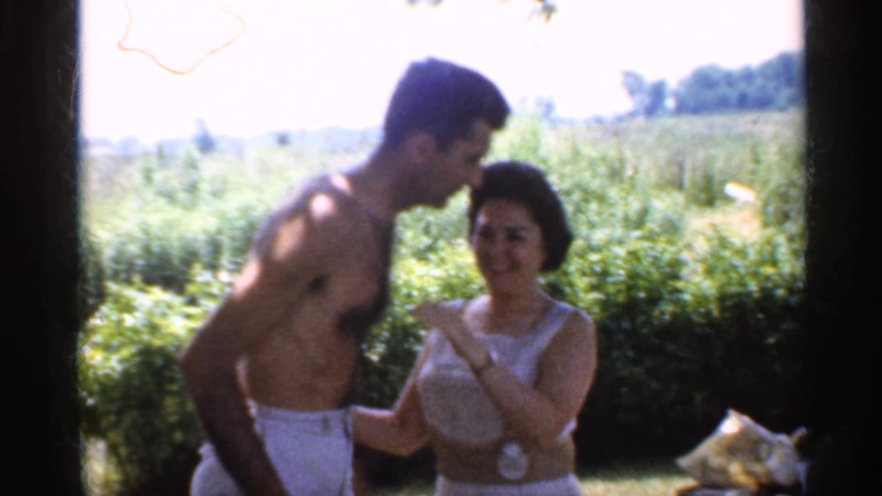 1959 Indiana backyard frame of a shirtless man leaning in to kiss or greet a smiling woman in a white dress.