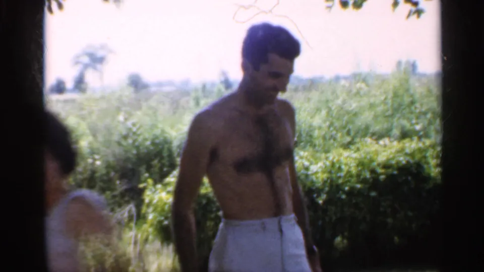 1959 Indiana home-movie frame of a shirtless man walking in a sunlit backyard while a woman is partly visible nearby.