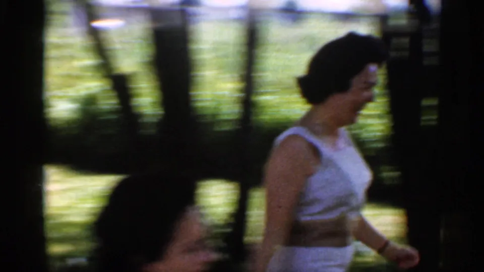 1959 Indiana frame of a woman in a white sleeveless dress moving through a shaded backyard with trees and lawn behind her.