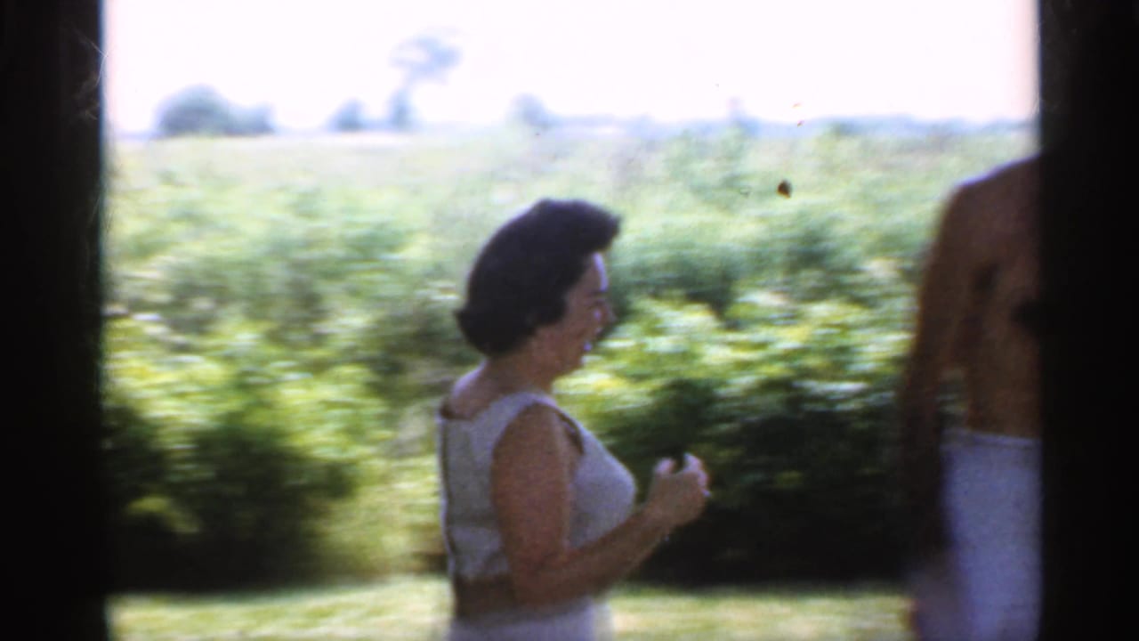 Woman in white sleeveless clothing walks near a sunny yard with hedges and fields behind her.