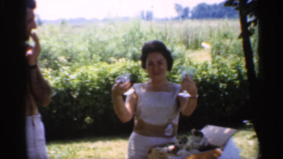 1959 Indiana 8mm frame showing a woman in white holding two small glasses while standing near a picnic basket outdoors.