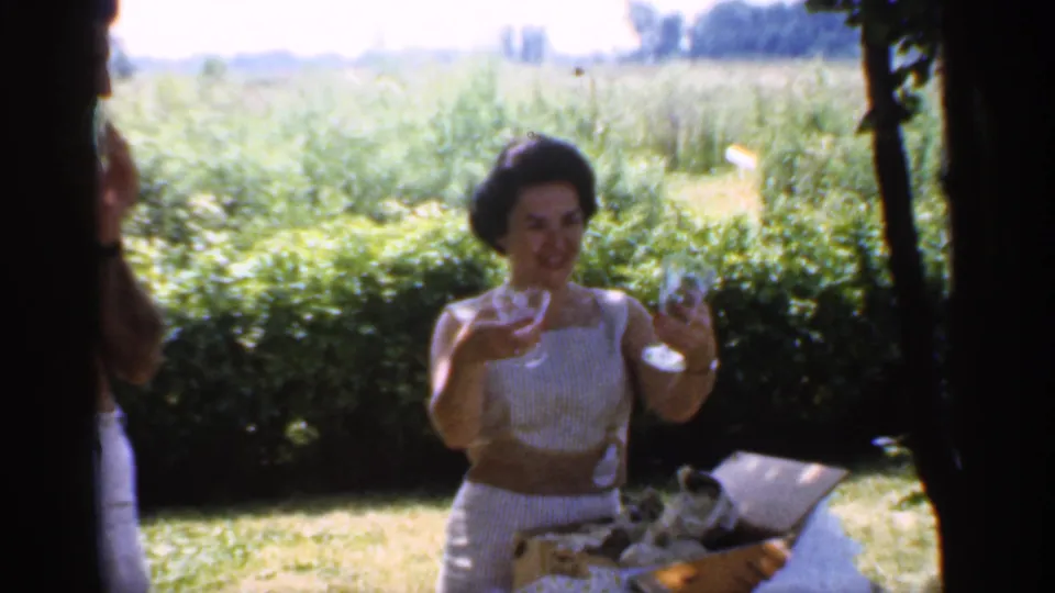 1959 Indiana home-movie frame with a woman smiling and holding small glasses near a picnic basket, hedges, and open fields.
