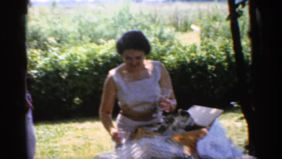 In Indiana home-movie footage from 1959, a woman in a white sleeveless dress sits near wrapped gifts on a grassy lawn framed by trees.