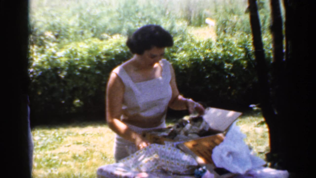 The 1959 Indiana home movie shows the woman looking down as she handles birthday gifts or wrapping beside grass and bushes.