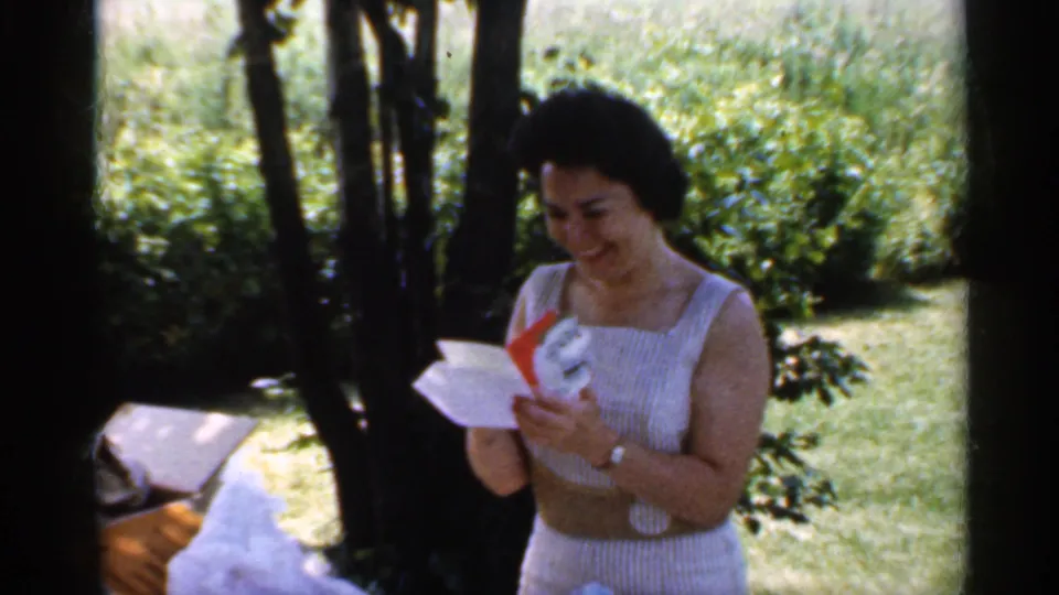 A smiling woman at the Indiana birthday picnic holds a card or paper, with wrapping and a bench or table visible beside her.