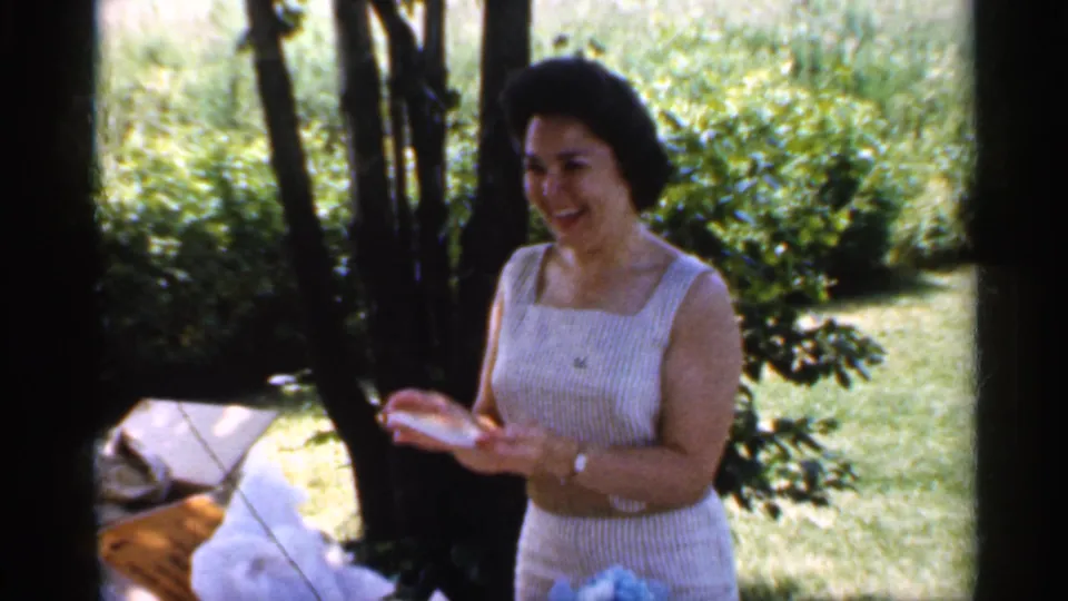 The woman smiles while holding a small light-colored gift at the shaded picnic setting, with lawn, bushes, and wrapping nearby.