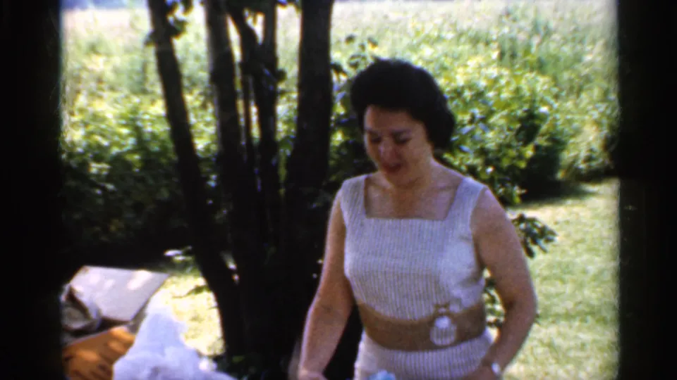 In 1959 Indiana home-movie footage, a woman in a white striped dress stands beside a table under trees in a garden.
