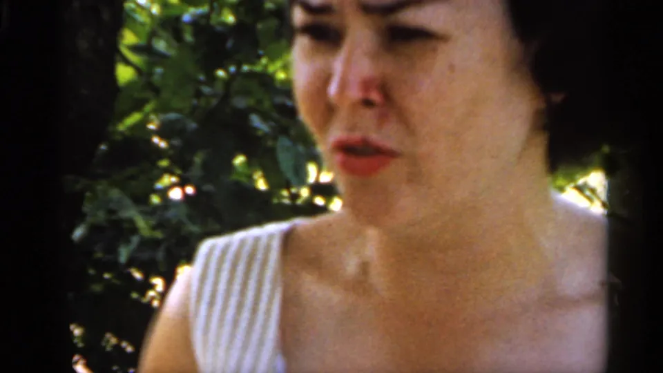 Close-up of the woman’s face and striped dress shoulder in soft 8mm focus, with leafy garden shade behind her.